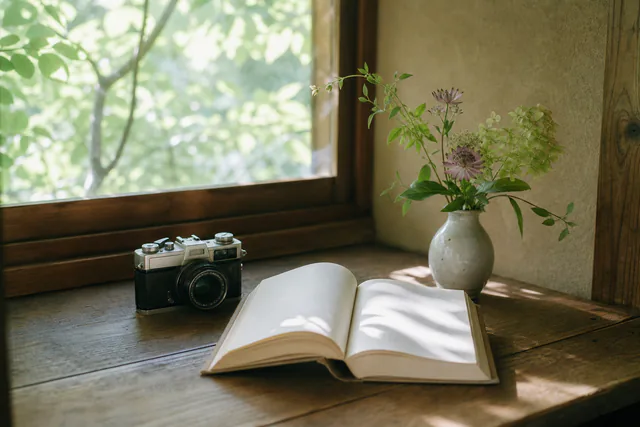 窓辺のフィルムカメラと読みかけの本、小さな花瓶の花